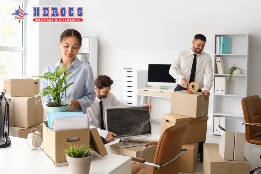 Office team packing boxes and organizing items during a multi location business move. Heroes Moving & Storage logo in top left corner.