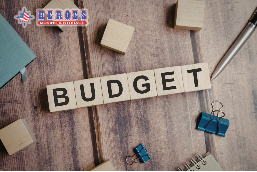 Wooden blocks spelling budget next to office supplies representing moving cost planning. The Heroes Moving & Storage logo is in the top left corner.