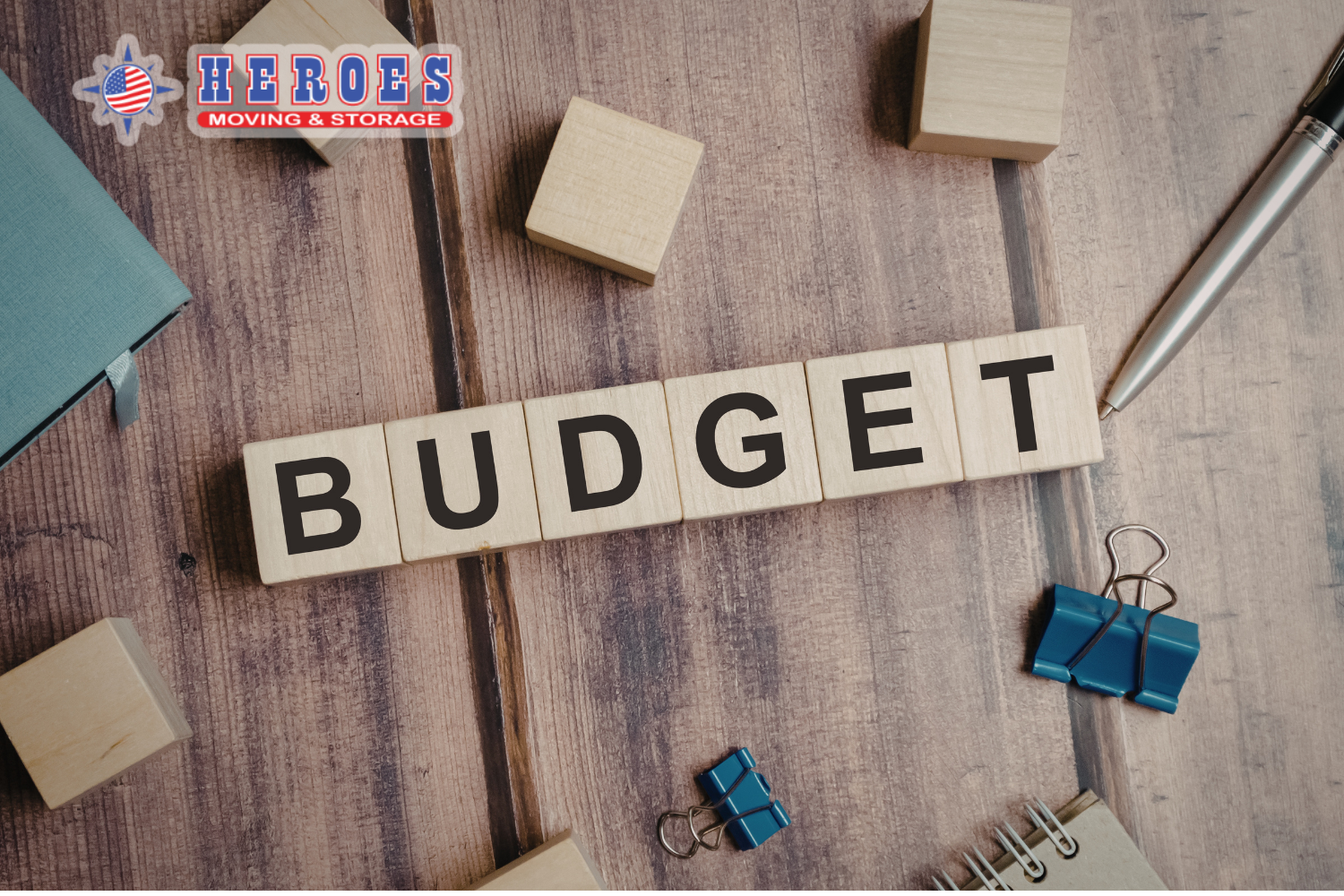 Wooden blocks spelling budget next to office supplies representing moving cost planning. The Heroes Moving & Storage logo is in the top left corner.