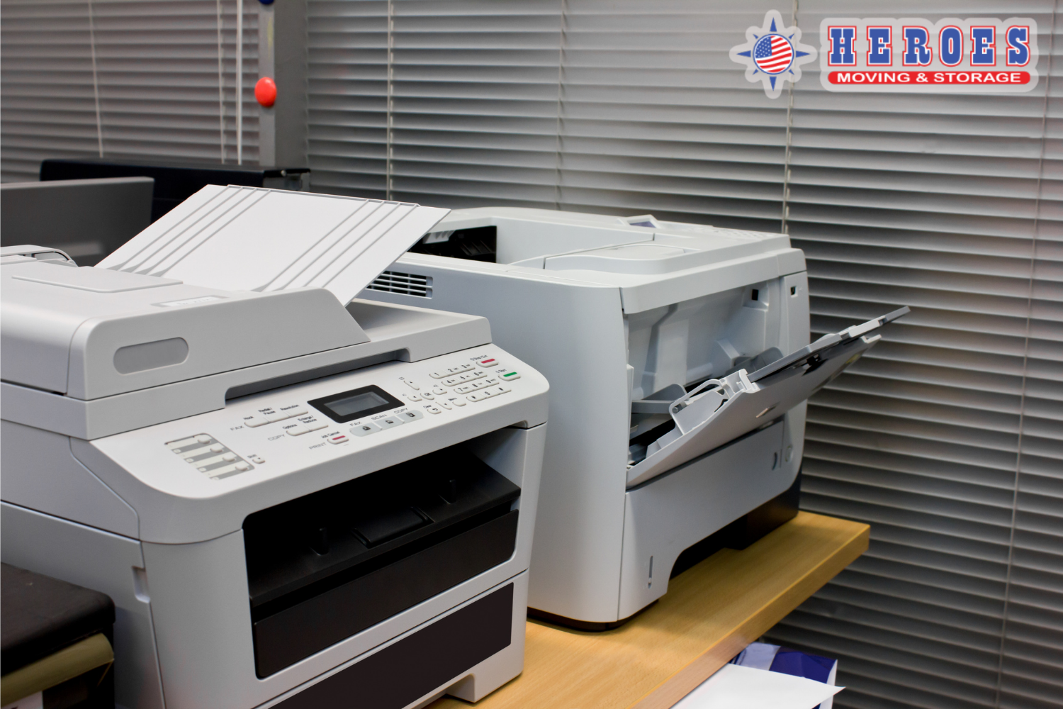 Office copier and printer equipment set up in a workspace during relocation. The Heroes Moving & Storage logo is in the top right corner.
