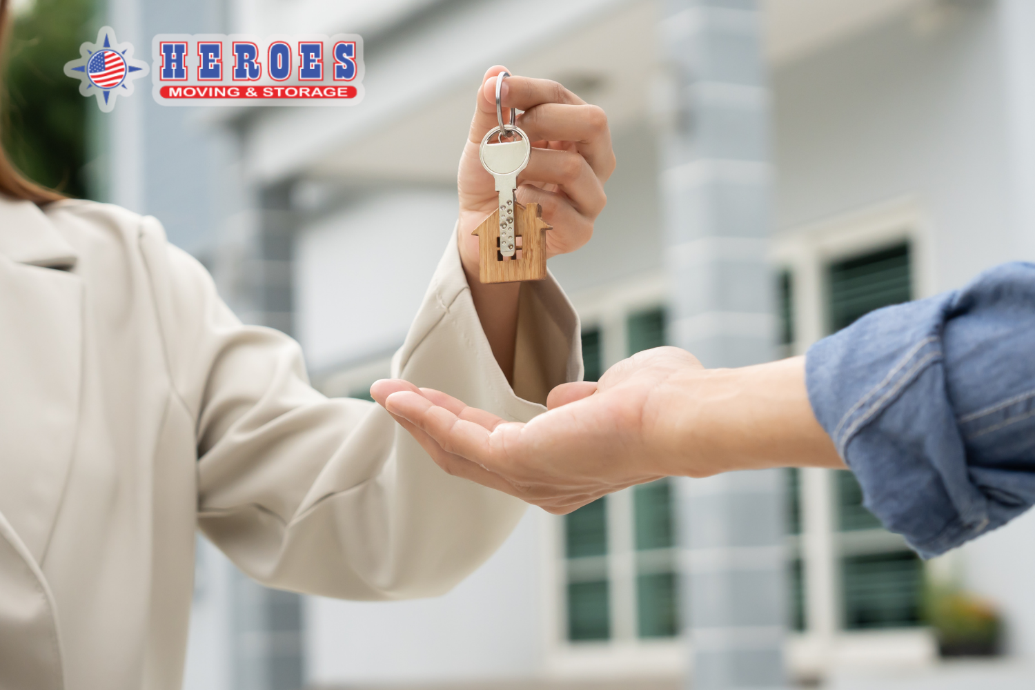 A woman in a beige suit is handing a house key to a person in a denim long-sleeved shirt with the sleeves rolled up. The Heroes Moving & Storage logo is in the top left corner.