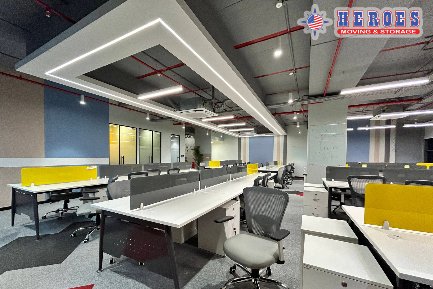 Modern office space with desks and chairs arranged for a business relocation walkthrough. The Heroes Moving & Storage logo is in the top right corner.